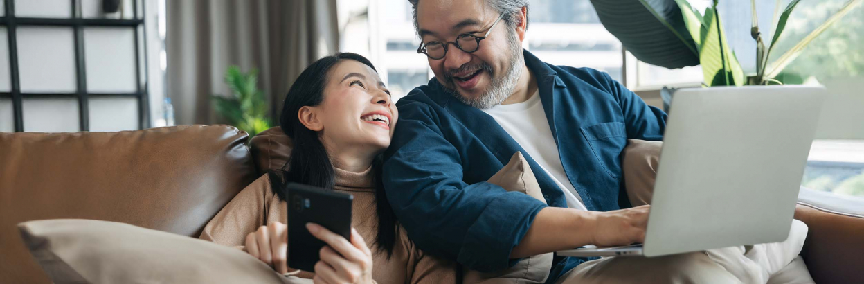 Two people sitting on couch smiling at each other while one uses cell phone and one uses laptop