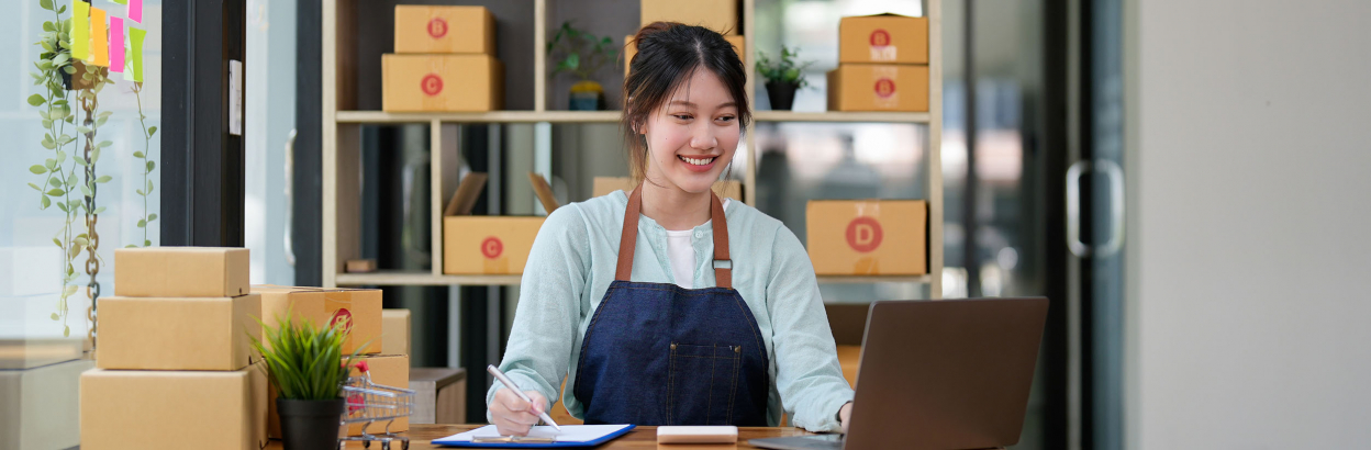 Female business owner using laptop at desk in her storefront with packages on a shelf behind her