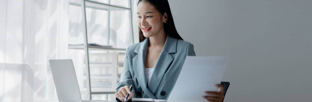Businesswoman working on laptop in office, holding paperwork in hand