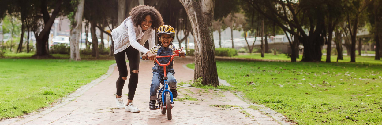 A mom pushing young child on a bike in a park on a sunny day