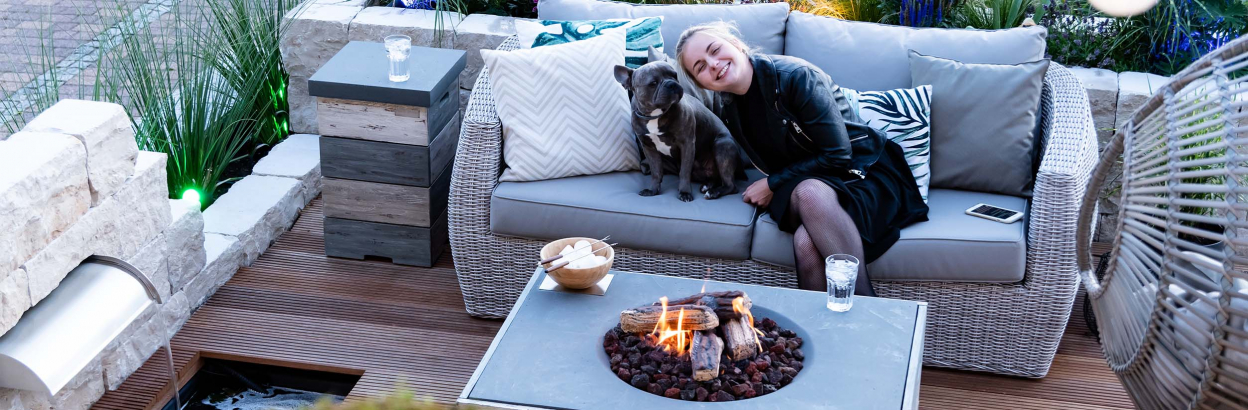 A young woman sitting outside on patio deck with French bulldog near a fire pit